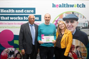 Three people look at the camera, standing in front of a Healthwatch sign