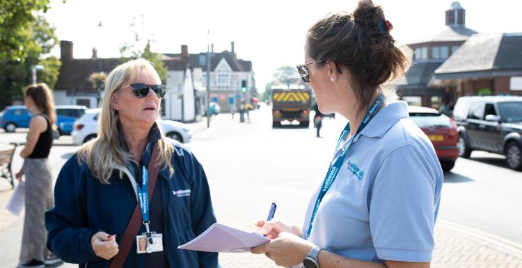 Two women in Healthwatch branded clothing are standing on a high street having a conversation.