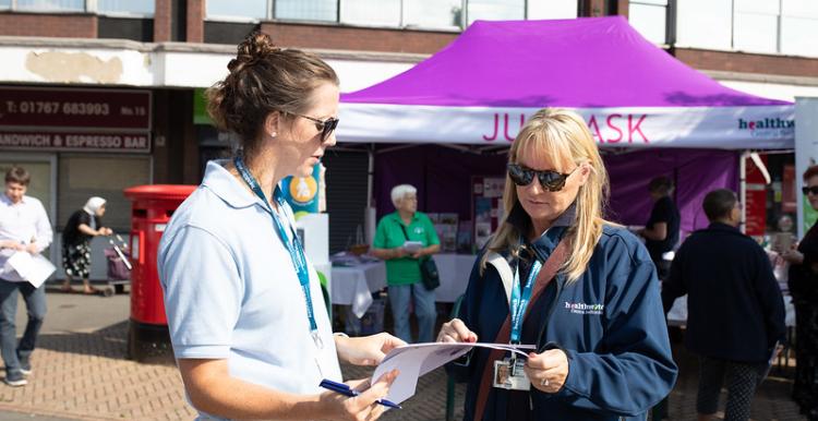 A woman with brown hair and wearing a light blue t-shirt is standing to the left. She is talking to a woman with blonde hair wearing a dark blue jacket. They are looking at a pamphlet. Both are wearing sunglasses.