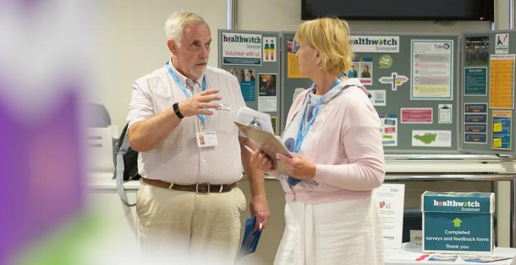 A man is standing on the left talking to a woman, standing on the right. The are standing in front of a Healthwatch information board.