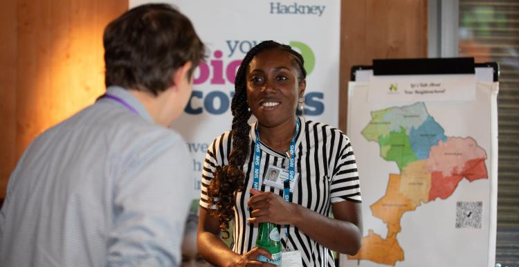 A male and female in conversation at an indoor community event. His back is to the camera, she is smiling at the camera. A Healthwatch banner and map of the local area are in the background.