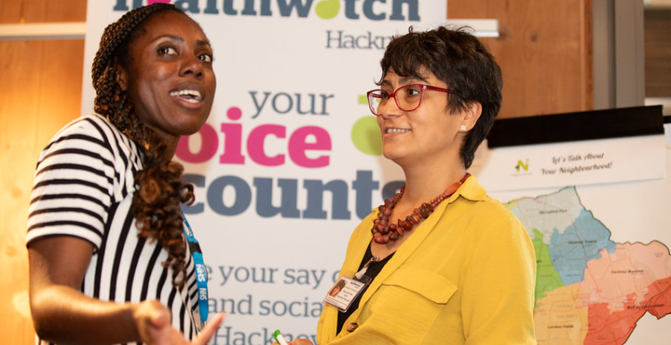 Two women from Healthwatch Hackney stand in front of a Healthwatch Hackney sign and map of Hackney. One is wearing a striped top, the other a yellow blouse and red glasses.