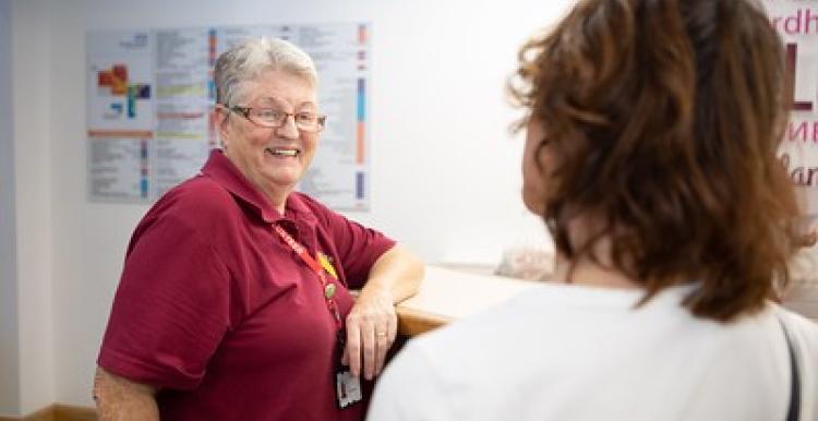 An older female Healthwatch volunteer smiles at a patient in a hospital setting