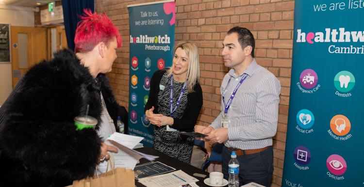 A woman with short red hair talking to a man and a woman behind a Healthwatch stall, at a community event.