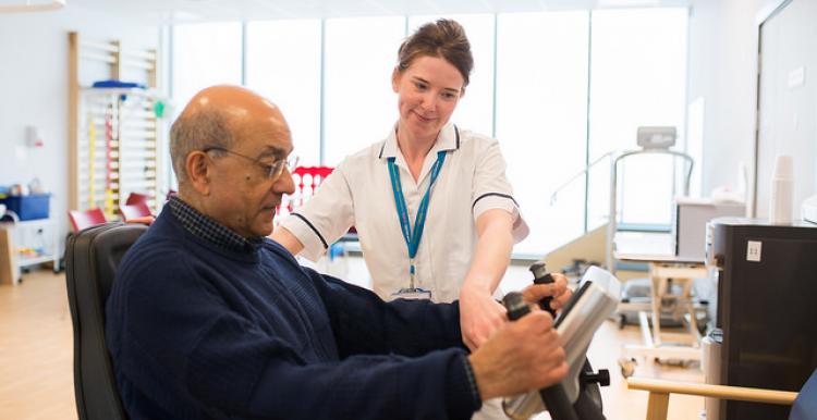 An older man in a blue jumper, being shown how to use a machine by a young female healthcare professional.