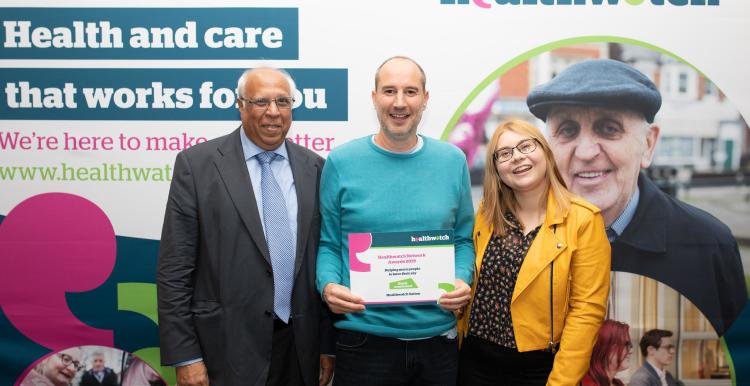 Three people look at the camera, standing in front of a Healthwatch sign