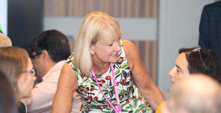 A woman with blonde hair, wearing a patterned dress, talking to someone at a table