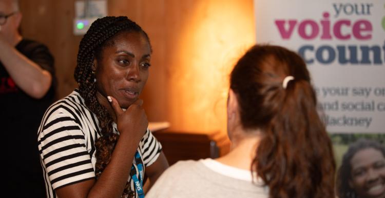 A young black woman in a black and white striped top is speaking with another person, they are defocused.