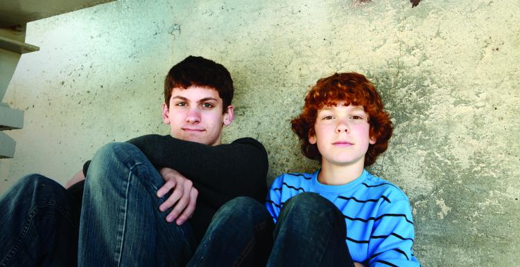 Two boys, sitting on the floor against a wall, smiling at the camera