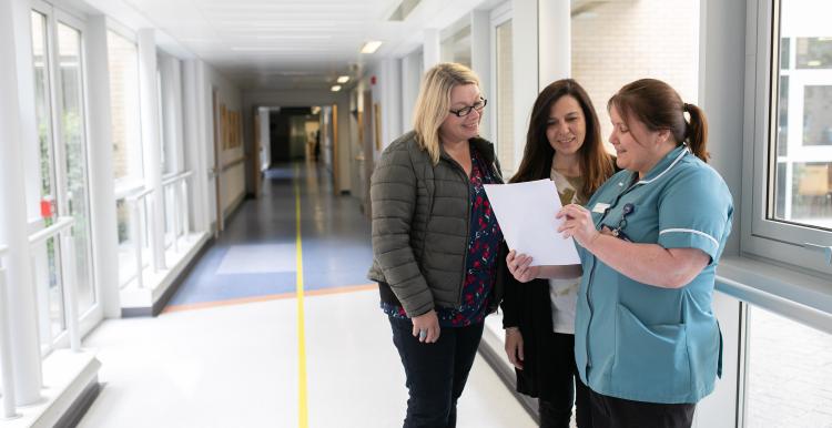 Three women including a clinician are standing in a hospital corridor reading a document.