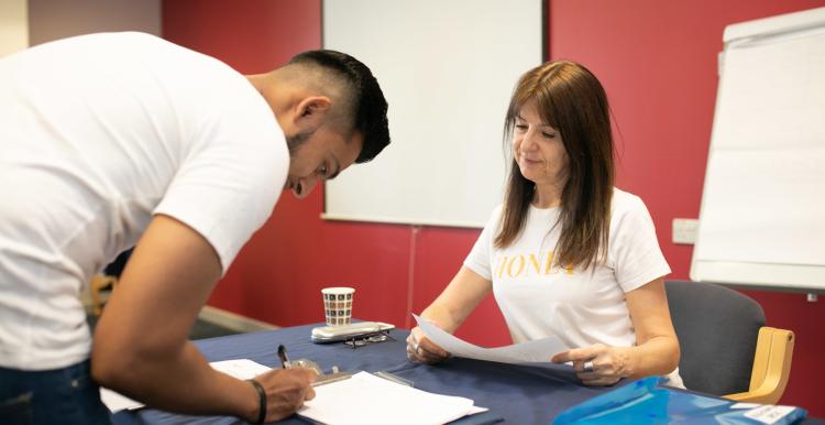 A young man in a white t-shirt is bent over a desk. He is signing a form. A woman is sitting at the desk. She is smiling.