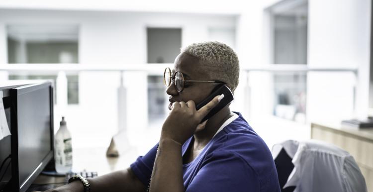 A person is sitting at a desk and computer. They have a phone to their ear, in their left hand. Their left side profile is visible. They are wearing a blue shirt.