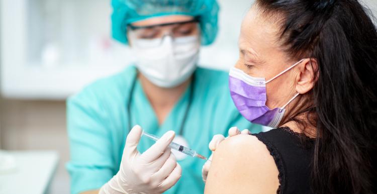 Nurse in protective work-wear holding syringe and injecting vaccine into senior woman's arm;