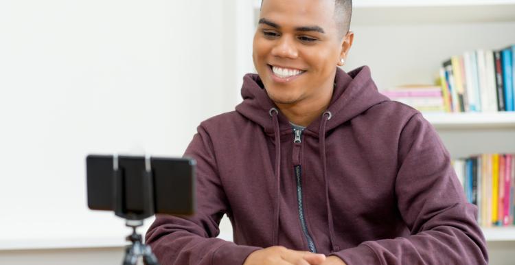 A male is smiling into a phone. The phone is in a tripod on a desk.