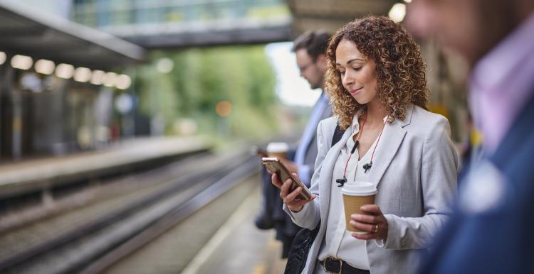 A woman standing on a train platform looking at her phone. She holds a disposable coffee cup in one hand.