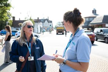 Two women standing outside on a sunny day. They are talking. One is holding a pen and paper. They are both wearing Healthwatch branded clothing.