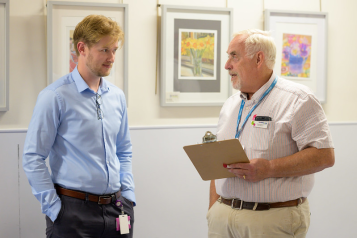 Two men standing in a clinical setting. On the left is a man wearing a blue shirt. On the white is a man with a cliipboard and lanyard.