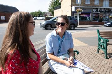 Two women sitting on a bench. They are in a town centre on a sunny day. The woman on the left is wearing a red top. The woman on the right is smiling, wearing sunglasses and wearing a light blue top with Healthwatch logo.