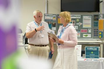 A man is standing on the left talking to a woman, standing on the right. The are standing in front of a Healthwatch information board.
