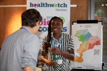 A male and female in conversation at an indoor community event. His back is to the camera, she is smiling at the camera. A Healthwatch banner and map of the local area are in the background.