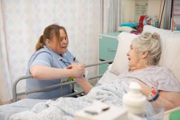 A female clinician sits next to a woman in a hospital bed. They are holding hands.