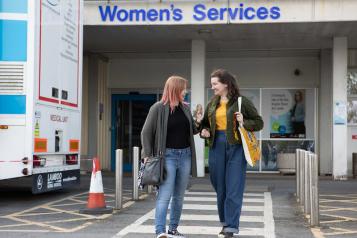 Two women walk out of a clinic. They are having a happy conversation. A sign behind them says 'women's services'.