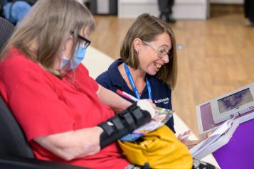 Woman in mask and wrist splint completing a form. A woman in medical uniform kneeling next to her and smiling