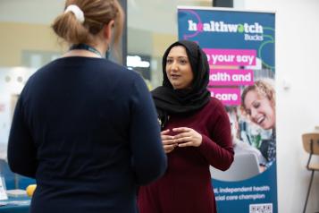 Two women talking in front of a Healthwatch pop up banner.  One is facing the camera.  One has her back to it. 