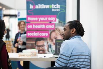 Woman standing next to Healthwatch pop up banner.  Man and woman talking at a table. 