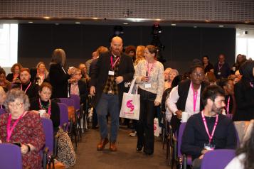 Lots of people sitting in rows at Healthwatch conference. Man and woman walking up the aisle talking. 