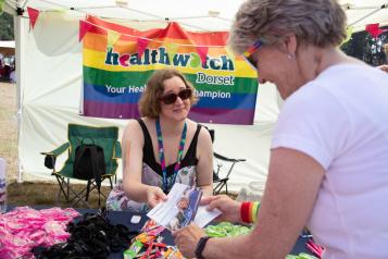 Woman handing a leaflet from a table in front of a rainbow Healthwatch banner to another woman opposite her