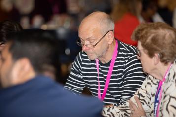 Man at table in glasses reading. Woman facing him and another man with back to the camera. 