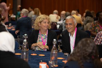 Two people at a table looking at the camera with lots of people behind them in a meeting room.
