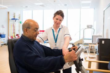 An older man in a blue jumper, being shown how to use a machine by a young female healthcare professional.