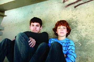 Two boys, sitting on the floor against a wall, smiling at the camera