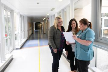 Three women including a clinician are standing in a hospital corridor reading a document.