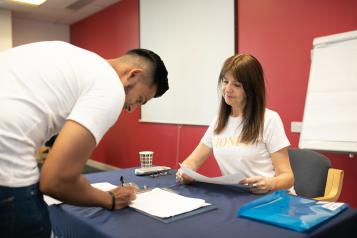 A young man in a white t-shirt is bent over a desk. He is signing a form. A woman is sitting at the desk. She is smiling.