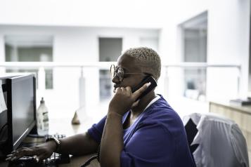 A person is sitting at a desk and computer. They have a phone to their ear, in their left hand. Their left side profile is visible. They are wearing a blue shirt.