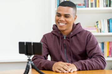 A male is smiling into a phone. The phone is in a tripod on a desk.