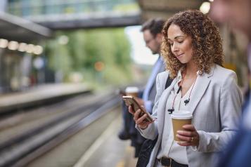 A woman standing on a train platform looking at her phone. She holds a disposable coffee cup in one hand.
