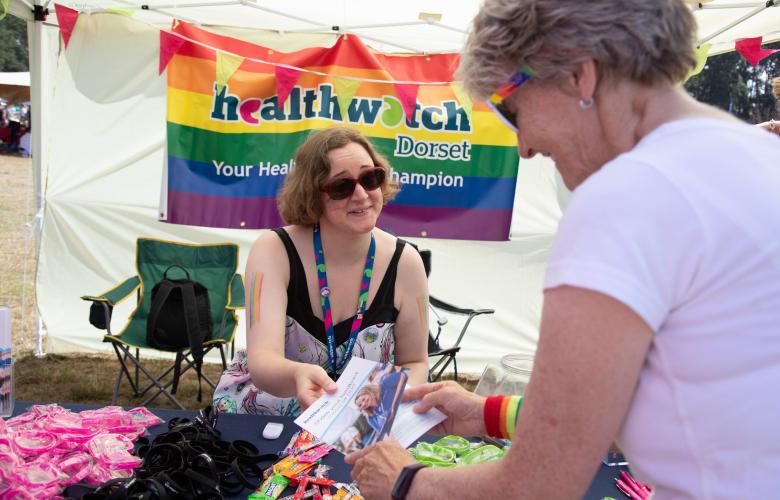 Woman handing a leaflet from a table in front of a rainbow Healthwatch banner to another woman opposite her